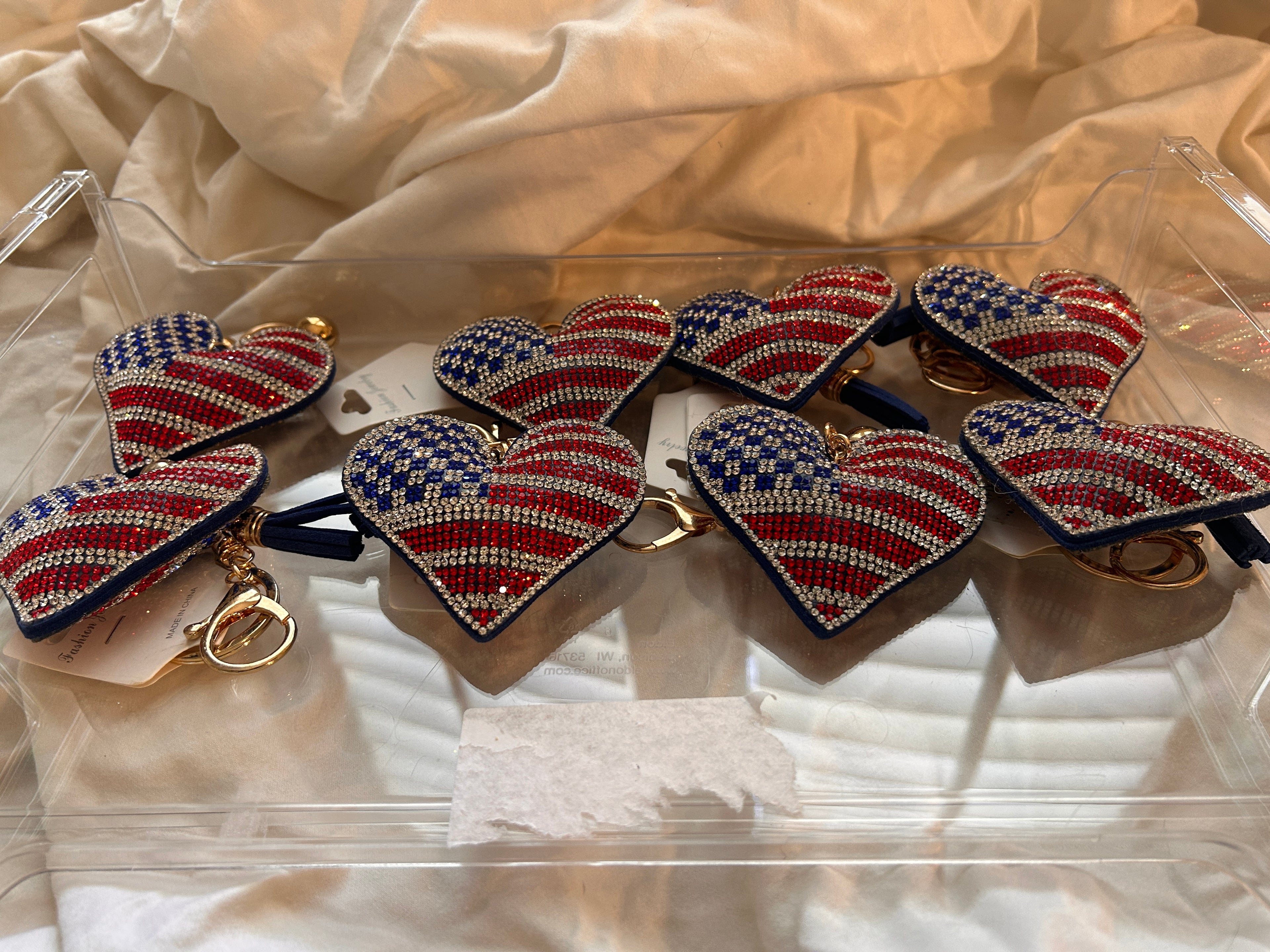 This Wrapables heart keychain features a puffy design adorned with red, white, and blue rhinestones forming an American flag pattern.It includes a gold-toned key ring, a lobster clasp for attachment to purses or handbags, and a dark blue tassel.The heart charm measures approximately 2.5 inches by 2.5 inches.The reverse side is made of a soft plush velvet material.