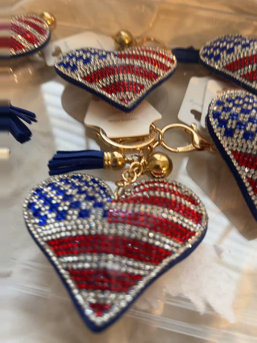 This Wrapables heart keychain features a puffy design adorned with red, white, and blue rhinestones forming an American flag pattern.It includes a gold-toned key ring, a lobster clasp for attachment to purses or handbags, and a dark blue tassel.The heart charm measures approximately 2.5 inches by 2.5 inches.The reverse side is made of a soft plush velvet material.