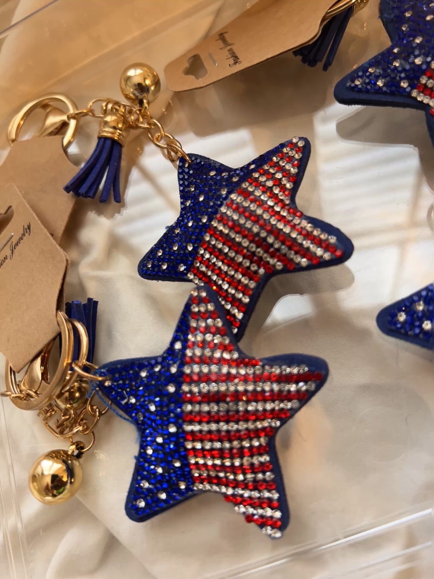 This Wrapables star keychain features a puffy design adorned with red, white, and blue rhinestones forming an American flag pattern.It includes a gold-toned key ring, a lobster clasp for attachment to purses or handbags, and a dark blue tassel.The star charm measures approximately 2.5 inches by 2.5 inches.The reverse side is made of a soft plush velvet material.
