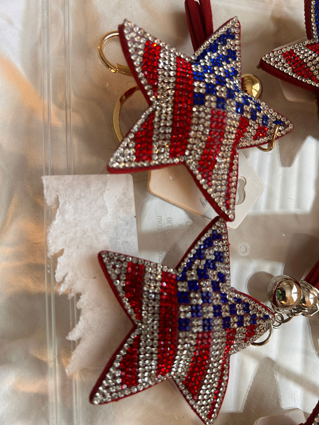 This Wrapables star keychain features a puffy design adorned with red, white, and blue rhinestones forming an American flag pattern.It includes a gold-toned key ring, a lobster clasp for attachment to purses or handbags, and a dark blue tassel.The star charm measures approximately 2.5 inches by 2.5 inches.The reverse side is made of a soft plush velvet material.